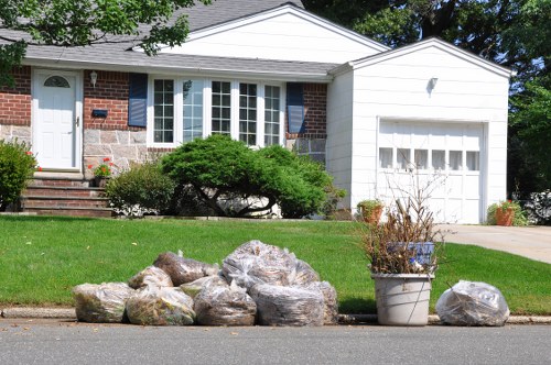 In front of a suburban house with a brick and white siding exterior, a collection of large, irregularly shaped rocks with textured surfaces and earthy tones are placed on the curbside. To the right, a white metal bucket containing dried, withered plants is positioned on the pavement. The house features a well-maintained green lawn, a shrubbery with dense foliage near the bay window, and a concrete path leading to the front door accessible via brick steps. A white garage door with small window panels is visible to the right of the house. The scene is bathed in natural daylight, highlighting the clean, tidy appearance of the front yard, which is typical of rubbish removal activities carried out by services such as Commercial Waste Blackheath near the SE3 postcode area in Blackheath.