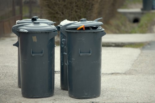 Two large, dark grey plastic wheelie bins with closed lids are positioned on a paved residential street, with one lid slightly ajar revealing an orange-handled tool or object inside. The bins are situated on the edge of a driveway or pavement, with a background of blurred greenery and a low stone wall, indicating a suburban or urban setting. The bins appear to be used for rubbish or general waste collection, commonly handled by rubbish removal services such as those offered by Commercial Waste Blackheath. The scene is illuminated by natural daylight, emphasizing the smooth, slightly reflective surface and rounded contours of the waste containers, which are standard in size and design for household refuse management in the local area around Blackheath, London.