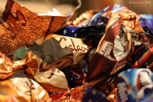 A close-up image showing a pile of crumpled, shiny foil wrappers and plastic packaging in various metallic and vibrant colors, including silver, copper, blue, and red, with some wrappers displaying partially visible text and brand logos. The wrappers are loosely arranged and overlapping, suggesting they are discarded packaging materials. The background is blurred but appears to be indoors with warm lighting, emphasizing the reflective surfaces and textured crumples of the wrappers. This image relates to waste materials that may be collected and removed by a rubbish collection service such as Commercial Waste Blackheath, serving the Blackheath area, which generally handles waste disposal and recycling of packaging debris.
