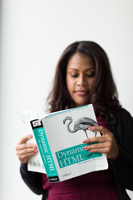 A woman with medium brown skin and long, dark hair is holding an open book titled 'Dynamic HTML' by O'Reilly, featuring a detailed illustration of a flamingo on the cover. She appears focused, with he